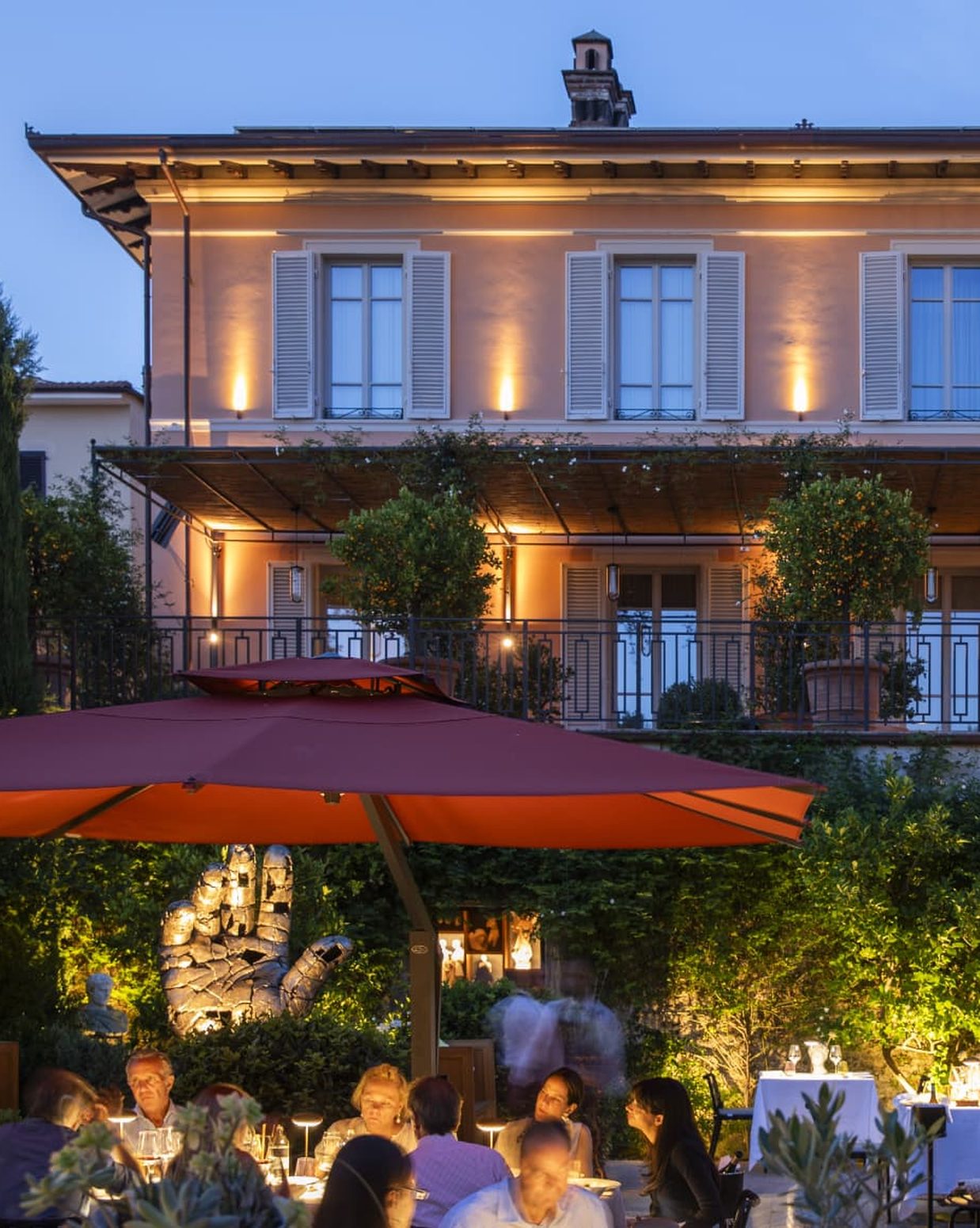 The garden terrace at Paradis Pietrasanta in the evening, with sculpture, a red parasol and warm light.